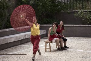 Interprète brandissant une ombrelle rouge, deux autres assises sur un banc dans une cour — Le bal des 3 p'tites têtes