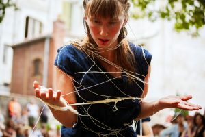 Danseuse enveloppée de fils de lin, mains ouvertes, devant une façade ensoleillée — Fibre