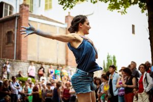 Danseuse de dos, bras tendu et main tachée de bleu, devant le public d'une place — Fibre