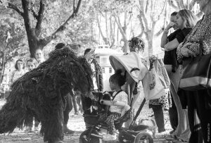 Silhouette en costume de fibres brunes penchée vers un enfant en tricycle, au milieu du public, en noir et blanc — Hors Sol