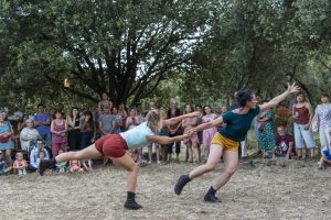 Deux danseuses s'élancent bras tendus l'une vers l'autre, public assis sous les chênes — Hors Sol